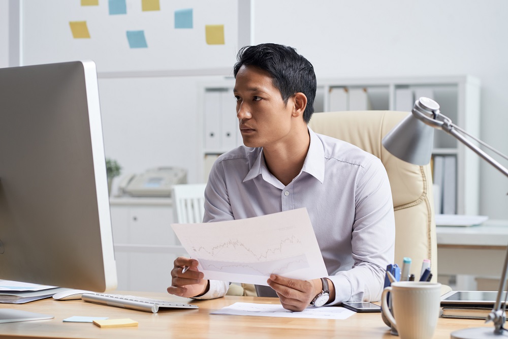 Financial analyst with document in his hands reading information on computer screen